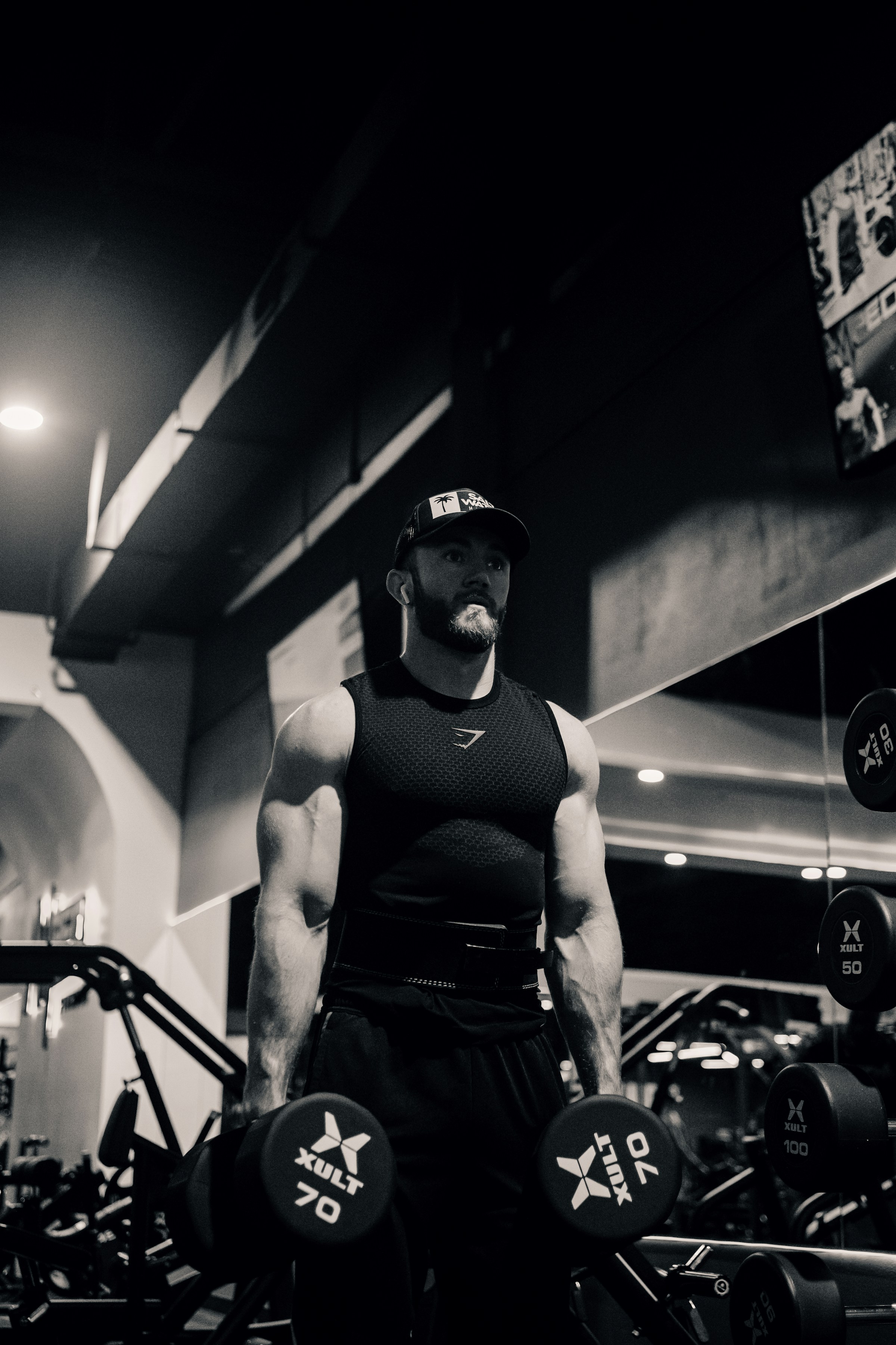 man in gym black and white pic holding dumbbells down at his sides ready to do dumbbell squat exercise in gym